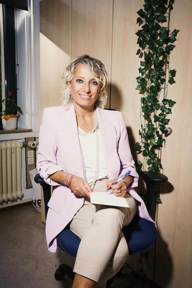 Foto: DIE KAVALLERIE GmbH Auf dem Foto ist eine sitzende Frau zu sehen, die einen Block und einen Stift in er Hand hält und in die Kamera lacht.