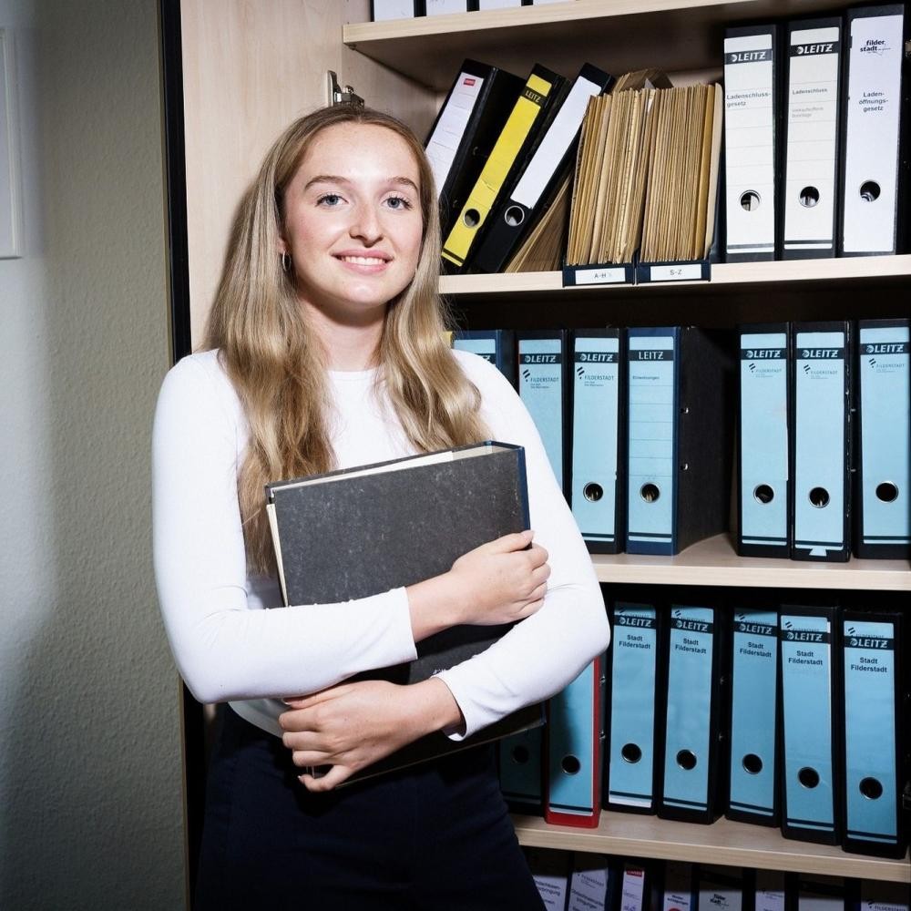 Foto: DIE KAVALLERIE GmbH Auf dem Foto ist einen Frau zu sehen, die vor einem Aktenschrank steht, einen Ordner in der Hand hält und in die Kamera schaut.