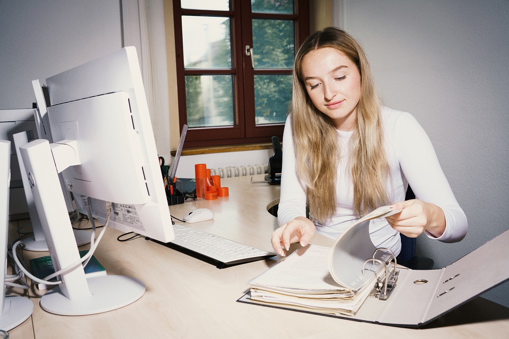 Auf dem Foto ist eine Frau an einem Schreibtisch zu sehen, die in einem Aktenordner blättert.