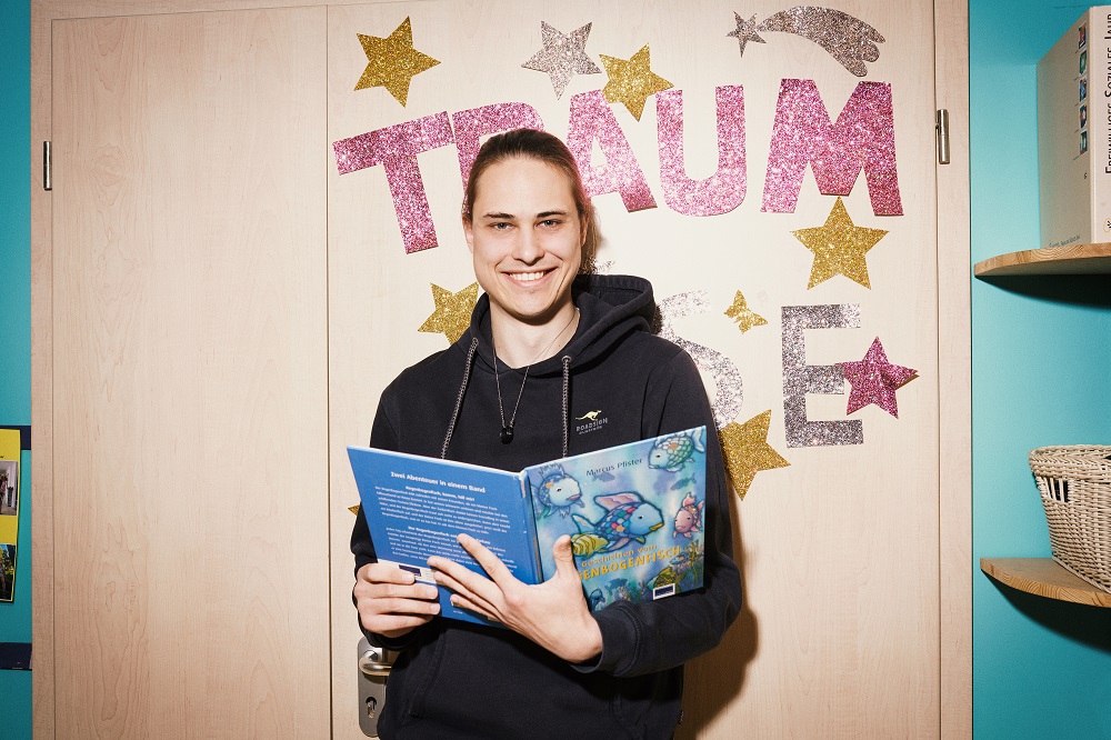 Auf dem Foto ist ein Mann zu sehen, der ein Kinderbuch in der Hand hält und in die Kamera lacht.