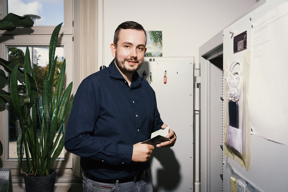 Auf dem Foto ist ein Mann zu sehen, der ein kleines Notizbuch in der Hand hält und in die Kamera lächelt. 